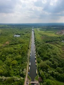Aerial view of a long canal with numerous boats surrounded by lush greenery and fields, under a partially cloudy sky.