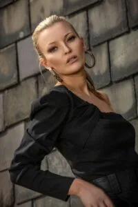 Woman in a black, one-shoulder top poses against a weathered brick wall, wearing large hoop earrings and a ponytail.