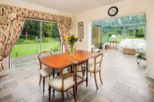 Dining room with wooden table, four chairs, and floral curtains, adjoining a sunlit conservatory with green garden view.