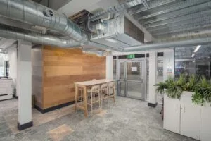 Modern office space with exposed ductwork, wooden wall paneling, a tall table with chairs, printer, exit door, and indoor plants.