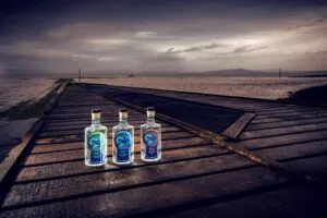 Three bottles of Stormy Bay gin on a weathered wooden pier with a stormy seascape in the background.