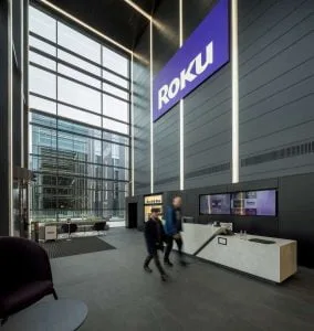 Modern office lobby with large "Roku" sign, reception desk, lounge seating, and two people walking past. Glass walls provide natural light.