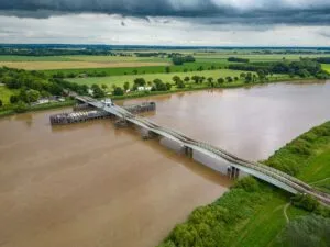 Aerial view of a partially constructed bridge over a wide river with surrounding green fields and overcast sky.